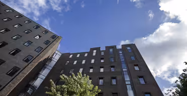 Blick von oben auf ein modernes Backsteinwohngebäude vor einem strahlend blauen Himmel mit vereinzelten Wolken. Im Vordergrund steht ein grüner Baum, der die unteren Gebäudeteile teilweise verdeckt.