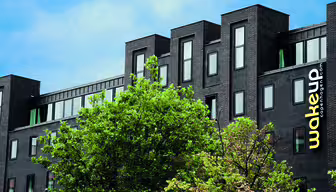 Modernes schwarzes Gebäude mit eleganten, rechteckigen Fenstern, umgeben von grünen Bäumen. Auf der Fassade prangt der gelbe Schriftzug „wakeup copenhagen.com“. Der Himmel ist teilweise bewölkt und blau.