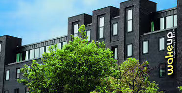 Modernes schwarzes Gebäude mit eleganten, rechteckigen Fenstern, umgeben von grünen Bäumen. Auf der Fassade prangt der gelbe Schriftzug „wakeup copenhagen.com“. Der Himmel ist teilweise bewölkt und blau.