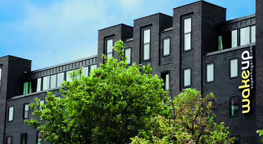 Modernes schwarzes Gebäude mit eleganten, rechteckigen Fenstern, umgeben von grünen Bäumen. Auf der Fassade prangt der gelbe Schriftzug „wakeup copenhagen.com“. Der Himmel ist teilweise bewölkt und blau.