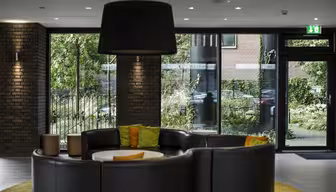 Moderne Bürolobby mit einem runden schwarzen Ledersofa, orangefarbenen und grünen Kissen und einem weißen runden Tisch in der Mitte. Große Fenster und Glastüren geben den Blick auf die grüne Umgebung frei, und über dem Sofa hängt eine große Deckenleuchte.