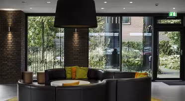 Moderne Bürolobby mit einem runden schwarzen Ledersofa, orangefarbenen und grünen Kissen und einem weißen runden Tisch in der Mitte. Große Fenster und Glastüren geben den Blick auf die grüne Umgebung frei, und über dem Sofa hängt eine große Deckenleuchte.