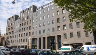Modernes, mehrstöckiges Backsteingebäude mit Fensterreihen und einem zentralen Glaseingang. Mehrere geparkte Autos säumen die Straße davor. Üppige grüne Bäume rahmen die Szene ein, und darüber ist ein blauer Himmel mit vereinzelten Wolken zu sehen.