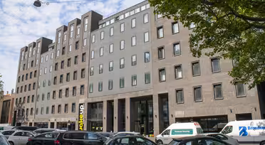 Modernes, mehrstöckiges Backsteingebäude mit Fensterreihen und einem zentralen Glaseingang. Mehrere geparkte Autos säumen die Straße davor. Üppige grüne Bäume rahmen die Szene ein, und darüber ist ein blauer Himmel mit vereinzelten Wolken zu sehen.