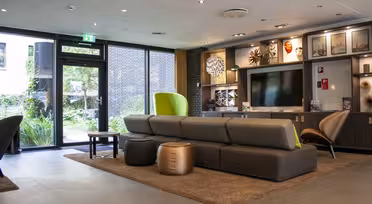 Ein moderner Wohnbereich mit einem großen grauen Sofa, braunen Sitzkissen und einem Fernseher auf einem Holzschrank. Kunst und Dekoartikel sind in Regalen ausgestellt. Große Fenster links bieten einen Blick ins Grüne. Ein grüner Sessel setzt einen Farbtupfer.