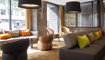 Moderner Loungebereich mit großen Fenstern, grauen Sofas, grünen und orangefarbenen Kissen, braunen Ledersesseln und schwarzen Pendelleuchten. Das einfallende Sonnenlicht schafft eine warme und einladende Atmosphäre.