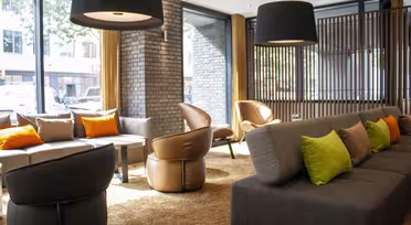 Moderner Loungebereich mit großen Fenstern, grauen Sofas, grünen und orangefarbenen Kissen, braunen Ledersesseln und schwarzen Pendelleuchten. Das einfallende Sonnenlicht schafft eine warme und einladende Atmosphäre.