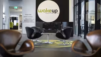 Moderne Hotellobby mit braunen Stühlen und kleinen Tischen auf einem grün-grauen Teppich. Ein großes Schild mit der Aufschrift „wakeupcopenhagen.com“ an einer dunklen Wand. Glastüren und ein Flur schaffen eine offene und einladende Atmosphäre.
