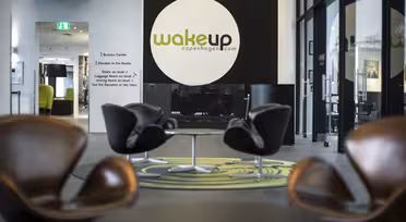 Moderne Hotellobby mit braunen Stühlen und kleinen Tischen auf einem grün-grauen Teppich. Ein großes Schild mit der Aufschrift „wakeupcopenhagen.com“ an einer dunklen Wand. Glastüren und ein Flur schaffen eine offene und einladende Atmosphäre.