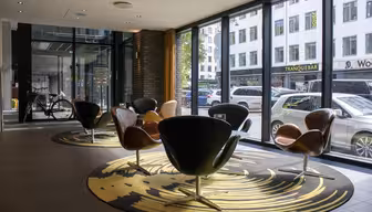 Moderne Lobby mit geschwungenen Stühlen auf einem runden Teppich vor raumhohen Fenstern. Draußen säumen geparkte Autos die Straße, im Hintergrund Geschäfte. In der Nähe des Eingangs ist ein Fahrrad zu sehen.