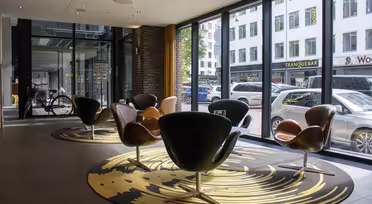 Moderne Lobby mit geschwungenen Stühlen auf einem runden Teppich vor raumhohen Fenstern. Draußen säumen geparkte Autos die Straße, im Hintergrund Geschäfte. In der Nähe des Eingangs ist ein Fahrrad zu sehen.
