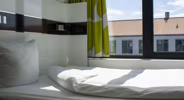 Ein ordentlich gemachtes Einzelbett mit weißer Bettwäsche und einem Kissen steht neben einem großen Fenster. Sonnenlicht strömt herein und erhellt den Raum. Grün-weiße Vorhänge hängen neben dem Fenster, das den Blick auf Gebäude mit roten Dächern freigibt.
