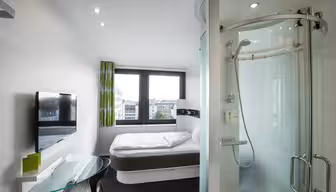 Modernes Hotelzimmer mit einem Doppelbett am Fenster, einer Glasduschkabine, einem an der Wand montierten Fernseher und einem kleinen Glasschreibtisch mit Stuhl. Das Zimmer hat weiße Wände, grüne Vorhänge und bietet Blick auf die Stadt.