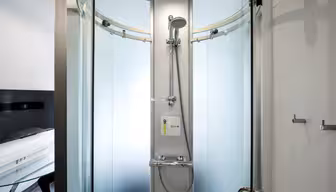 Eine moderne Duschkabine aus gebogenem Glas mit mattierten Paneelen. Sie verfügt über einen zentralen Duschkopf, eine Handbrause und ein kleines weißes Bedienfeld. An den Glastüren sind zwei Metallgriffe sichtbar. Der Raum besticht durch sein schlichtes, minimalistisches Design.