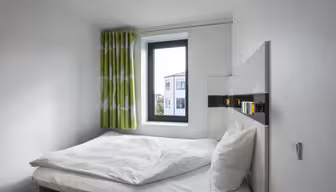 Ein kleines, modernes Schlafzimmer mit einem sauber gemachten Bett mit weißer Bettwäsche und Kissen. Ein Fenster mit grün-weißen Vorhängen lässt Tageslicht herein und gibt den Blick auf ein Gebäude frei. Das Zimmer ist minimalistisch eingerichtet und hat weiße Wände.