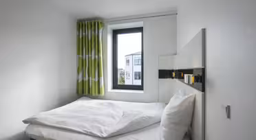 Ein kleines, modernes Schlafzimmer mit einem sauber gemachten Bett mit weißer Bettwäsche und Kissen. Ein Fenster mit grün-weißen Vorhängen lässt Tageslicht herein und gibt den Blick auf ein Gebäude frei. Das Zimmer ist minimalistisch eingerichtet und hat weiße Wände.