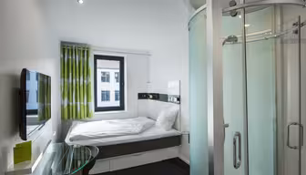 Kompaktes, modernes Hotelzimmer mit Bett, grün-weißen Vorhängen, Wandfernseher und einer Duschkabine aus gebogenem Glas. Neben dem Bett steht ein kleiner Glasschreibtisch. Das Fenster bietet Blick auf die Nachbargebäude.