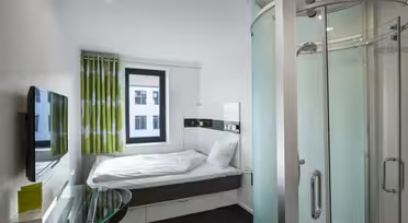 Kompaktes, modernes Hotelzimmer mit Bett, grün-weißen Vorhängen, Wandfernseher und einer Duschkabine aus gebogenem Glas. Neben dem Bett steht ein kleiner Glasschreibtisch. Das Fenster bietet Blick auf die Nachbargebäude.