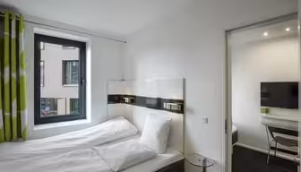 Ein modernes Hotelzimmer mit einem sauber gemachten Bett mit weißer Bettwäsche und einem grünen Vorhang. Ein Fenster bietet Blick auf das Nachbargebäude. Ein Flachbildfernseher und ein Schreibtisch sind durch eine offene Tür sichtbar, die in einen separaten Bereich führt.