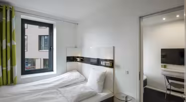 Ein modernes Hotelzimmer mit einem sauber gemachten Bett mit weißer Bettwäsche und einem grünen Vorhang. Ein Fenster bietet Blick auf das Nachbargebäude. Ein Flachbildfernseher und ein Schreibtisch sind durch eine offene Tür sichtbar, die in einen separaten Bereich führt.
