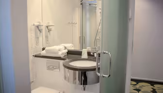 Modernes Badezimmer mit rundem Spiegel über einem weißen Waschbecken mit Chromarmatur. Handtücher liegen ordentlich auf der Ablage neben wandmontierten Haartrocknern. Im Hintergrund ist eine Glasduschkabine zu sehen. Der Boden ist mit gemusterten Fliesen ausgelegt.
