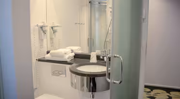 Modernes Badezimmer mit rundem Spiegel über einem weißen Waschbecken mit Chromarmatur. Handtücher liegen ordentlich auf der Ablage neben wandmontierten Haartrocknern. Im Hintergrund ist eine Glasduschkabine zu sehen. Der Boden ist mit gemusterten Fliesen ausgelegt.