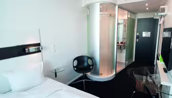 Modernes Hotelzimmer mit Einzelbett und elegantem schwarzen Sessel. In der Nähe des Eingangs befindet sich eine runde Glasdusche. Auf einem Glastisch steht ein Fernseher, daneben ein kleiner Arbeitsplatz. Der Innenraum ist hell mit weißen Wänden und minimalistischem Dekor.