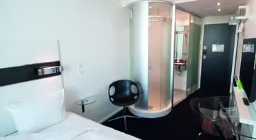 Modernes Hotelzimmer mit Einzelbett und elegantem schwarzen Sessel. In der Nähe des Eingangs befindet sich eine runde Glasdusche. Auf einem Glastisch steht ein Fernseher, daneben ein kleiner Arbeitsplatz. Der Innenraum ist hell mit weißen Wänden und minimalistischem Dekor.