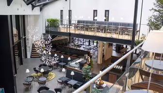 Modernes zweistöckiges Café mit Glasdach. Im Erdgeschoss sind runde Tische und Stühle großzügig angeordnet. Ein Balkon mit zusätzlichen Sitzgelegenheiten säumt die obere Ebene. Zwei Kronleuchter hängen, und Pflanzen sorgen für Grün im gesamten Raum.