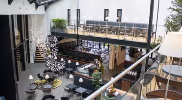 Modernes zweistöckiges Café mit Glasdach. Im Erdgeschoss sind runde Tische und Stühle großzügig angeordnet. Ein Balkon mit zusätzlichen Sitzgelegenheiten säumt die obere Ebene. Zwei Kronleuchter hängen, und Pflanzen sorgen für Grün im gesamten Raum.