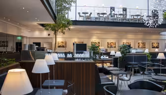 Modernes Restaurant-Interieur mit schwarzen Tischen und Stühlen, weißen Lampen und Topfbäumen. Ein Zwischengeschoss bietet zusätzliche Sitzplätze. Kunstwerke schmücken die Wände, und natürliches Licht durchflutet den großzügigen Raum.