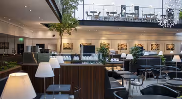 Modernes Restaurant-Interieur mit schwarzen Tischen und Stühlen, weißen Lampen und Topfbäumen. Ein Zwischengeschoss bietet zusätzliche Sitzplätze. Kunstwerke schmücken die Wände, und natürliches Licht durchflutet den großzügigen Raum.