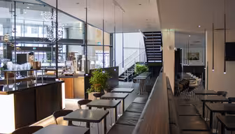 Modernes Café-Interieur mit eleganten schwarzen Tischen und Stühlen, großen Glasfenstern und minimalistischen Pendelleuchten. Eine Treppe führt nach oben, und Topfpflanzen sorgen für Grün. Helles Tageslicht durchflutet den Raum.