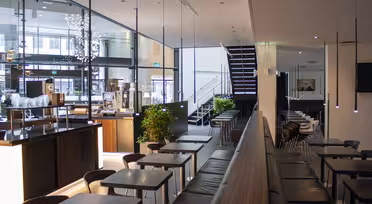 Modernes Café-Interieur mit eleganten schwarzen Tischen und Stühlen, großen Glasfenstern und minimalistischen Pendelleuchten. Eine Treppe führt nach oben, und Topfpflanzen sorgen für Grün. Helles Tageslicht durchflutet den Raum.