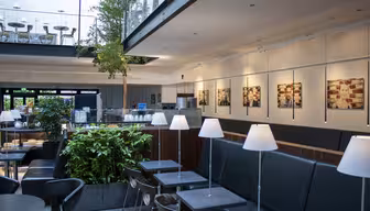 Modernes Café-Interieur mit schwarzen Sitzgelegenheiten, kleinen Tischen und weißen Lampen. Grüne Pflanzen sorgen für einen Hauch von Natur. Gerahmte Bilder säumen die Wand, darüber ist ein Zwischengeschoss mit Stühlen sichtbar. Helle und großzügige Atmosphäre.
