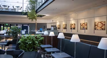 Modernes Café-Interieur mit schwarzen Sitzgelegenheiten, kleinen Tischen und weißen Lampen. Grüne Pflanzen sorgen für einen Hauch von Natur. Gerahmte Bilder säumen die Wand, darüber ist ein Zwischengeschoss mit Stühlen sichtbar. Helle und großzügige Atmosphäre.