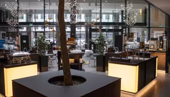 Ein modernes, offenes Café mit großen Glasfenstern und dekorativen Hängelampen. Ein Baum in einem Pflanzkübel steht zentral zwischen Holz- und Glasmöbeln. Im Hintergrund sind verschiedene Sitzbereiche zu sehen, in deren Nähe sich Kaffeemaschinen und Essensstände befinden.