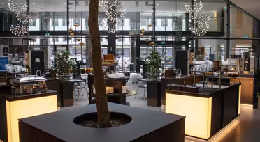 Ein modernes, offenes Café mit großen Glasfenstern und dekorativen Hängelampen. Ein Baum in einem Pflanzkübel steht zentral zwischen Holz- und Glasmöbeln. Im Hintergrund sind verschiedene Sitzbereiche zu sehen, in deren Nähe sich Kaffeemaschinen und Essensstände befinden.
