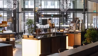 Modernes Cafeteria-Interieur mit eleganten Theken aus dunklem Holz, sanft beleuchtet. Eine Kaffeemaschine, Stapel weißer Teller und üppige grüne Pflanzen sind zu sehen. Große Fenster geben den Blick auf die Straße frei.