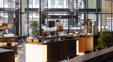 Modernes Cafeteria-Interieur mit eleganten Theken aus dunklem Holz, sanft beleuchtet. Eine Kaffeemaschine, Stapel weißer Teller und üppige grüne Pflanzen sind zu sehen. Große Fenster geben den Blick auf die Straße frei.