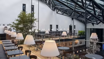 Modernes Café mit hoher Glasdecke und schwarzen Metallträgern. Ein großer Baum, Holzboden, Lampen auf den Tischen und Sitzbereiche auf zwei Ebenen schaffen eine luftige und moderne Atmosphäre.