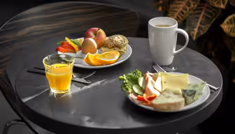 Ein kleiner runder Tisch mit zwei Tellern mit Frühstücksgerichten. Auf einem Teller liegen ein Bagel, ein Ei, Orangenscheiben und ein Pfirsich. Auf dem anderen liegen Brot, Gemüse und Käse. Auf dem Tisch stehen ein Glas Orangensaft und eine weiße Tasse.