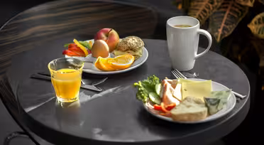 Ein kleiner runder Tisch mit zwei Tellern mit Frühstücksgerichten. Auf einem Teller liegen ein Bagel, ein Ei, Orangenscheiben und ein Pfirsich. Auf dem anderen liegen Brot, Gemüse und Käse. Auf dem Tisch stehen ein Glas Orangensaft und eine weiße Tasse.