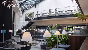 Modernes Café-Interieur mit großen Fenstern, freiliegenden Balken und stilvollen Hängelampen. Schwarze Tische und Stühle verteilen sich auf zwei Ebenen, eine Treppe führt zum oberen Essbereich. Grüne Pflanzen sorgen für einen Hauch von Natur.