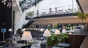 Modernes Café-Interieur mit großen Fenstern, freiliegenden Balken und stilvollen Hängelampen. Schwarze Tische und Stühle verteilen sich auf zwei Ebenen, eine Treppe führt zum oberen Essbereich. Grüne Pflanzen sorgen für einen Hauch von Natur.