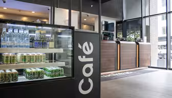 Café-Interieur mit einer Glasvitrine, in der Wasserflaschen und Getränkedosen präsentiert werden. Im Hintergrund befindet sich eine moderne Theke mit Holzpaneelen, während große Fenster den Blick nach draußen ermöglichen. Der Raum ist elegant und modern gestaltet.