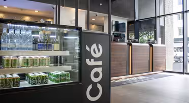 Café-Interieur mit einer Glasvitrine, in der Wasserflaschen und Getränkedosen präsentiert werden. Im Hintergrund befindet sich eine moderne Theke mit Holzpaneelen, während große Fenster den Blick nach draußen ermöglichen. Der Raum ist elegant und modern gestaltet.
