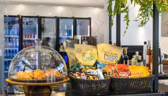 Eine Café-Theke mit einer Glaskuppel, Croissants und Körben mit verschiedenen Snacks wie Chips, Nüssen und Süßigkeiten. Dahinter befinden sich Glaskühlschränke mit verschiedenen Getränken. Hängepflanzen schmücken den Bereich.