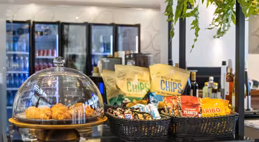 Eine Café-Theke mit einer Glaskuppel, Croissants und Körben mit verschiedenen Snacks wie Chips, Nüssen und Süßigkeiten. Dahinter befinden sich Glaskühlschränke mit verschiedenen Getränken. Hängepflanzen schmücken den Bereich.