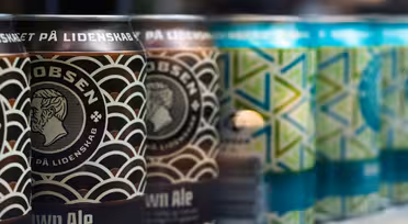 Eine Reihe von Getränkedosen mit aufwendigen Designs und Mustern. Die nächste Dose trägt ein kreisförmiges Emblem mit Text und geometrischen Motiven, während die anderen blaue und grüne geometrische Formen zeigen. Der Fokus liegt auf den künstlerischen Details.